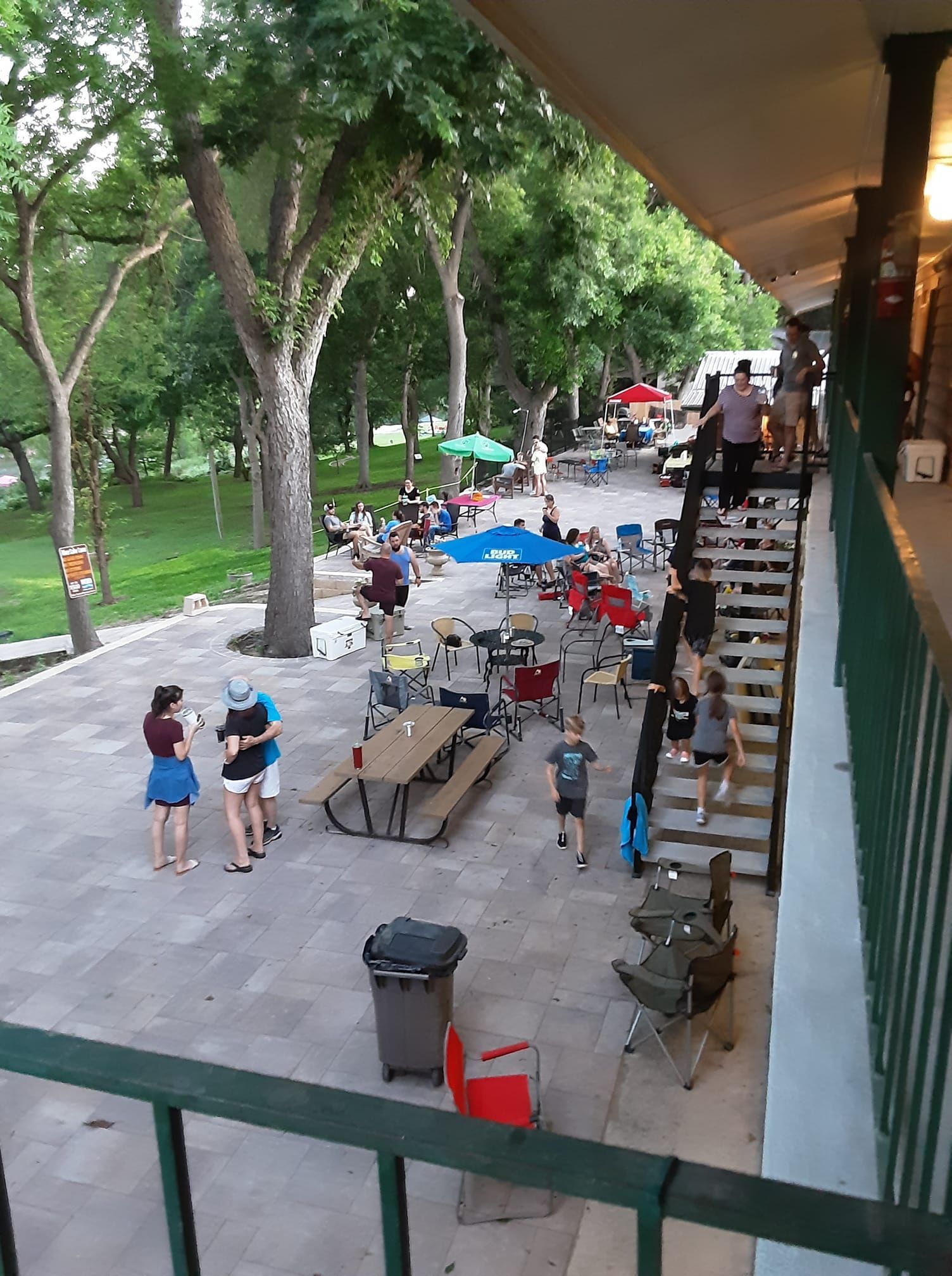 People gathered outdoors at a patio with tables and chairs, trees, and a building with stairs; daytime.