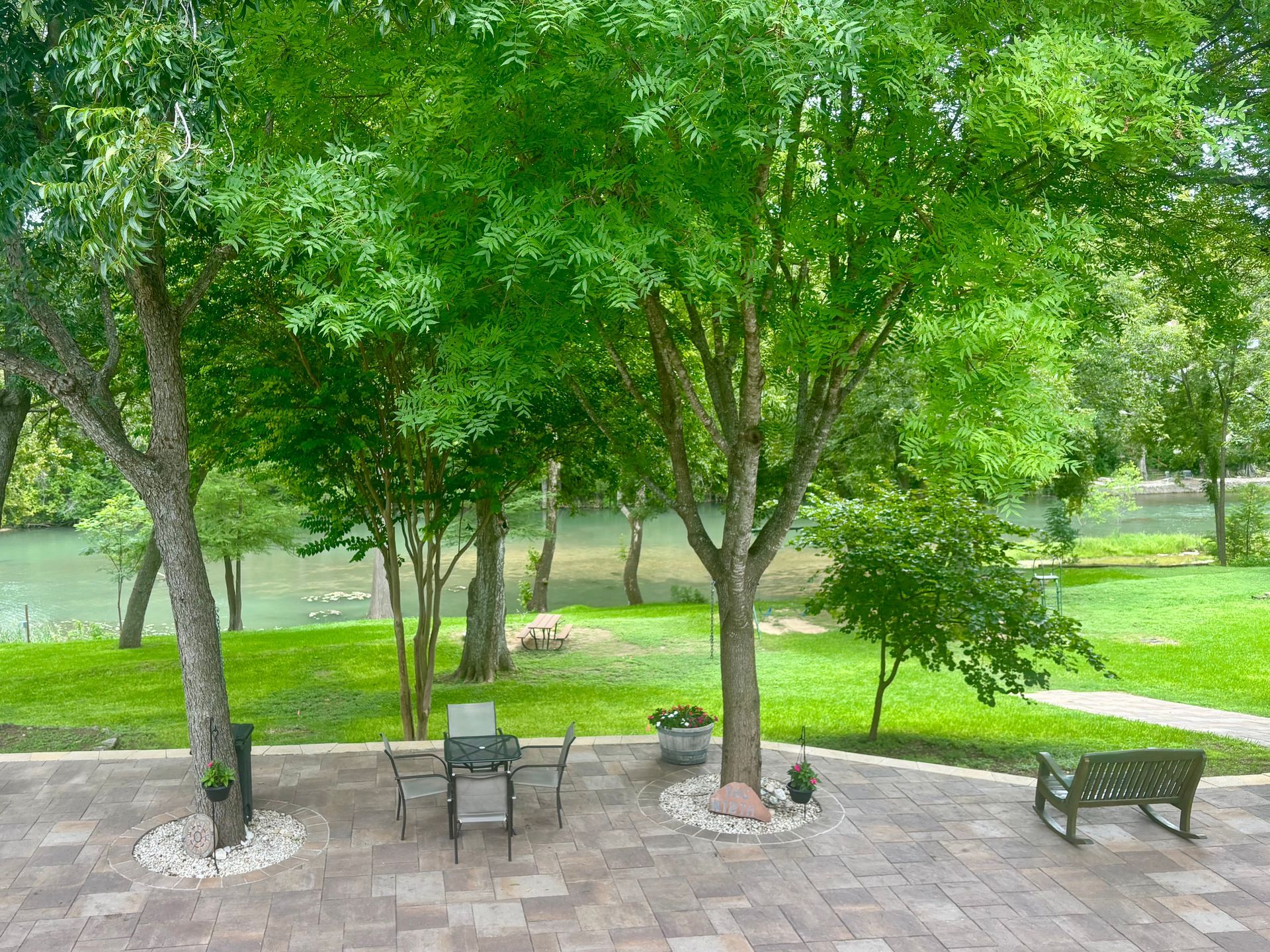 Patio with table and chairs under green trees, overlooking a river and lawn.
