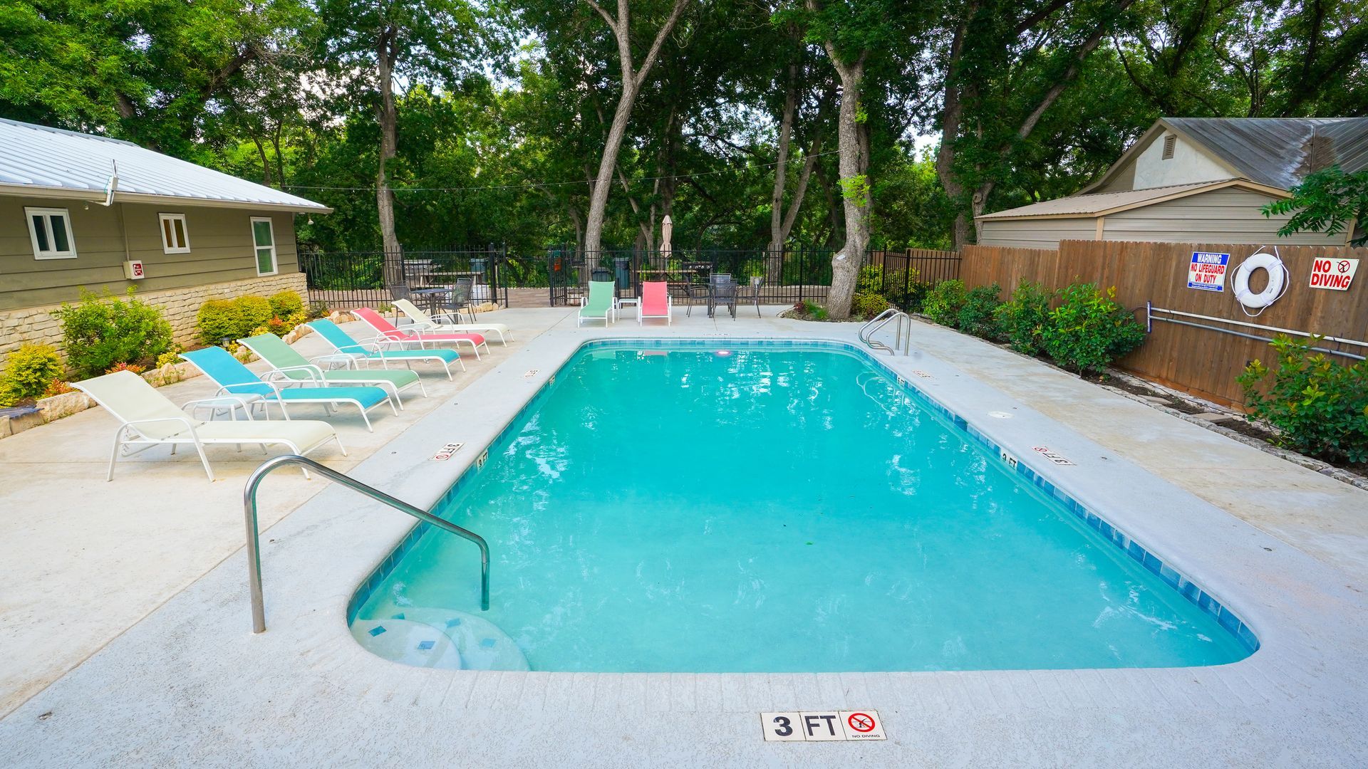 Swimming pool surrounded by lounge chairs; a building and trees in the background.
