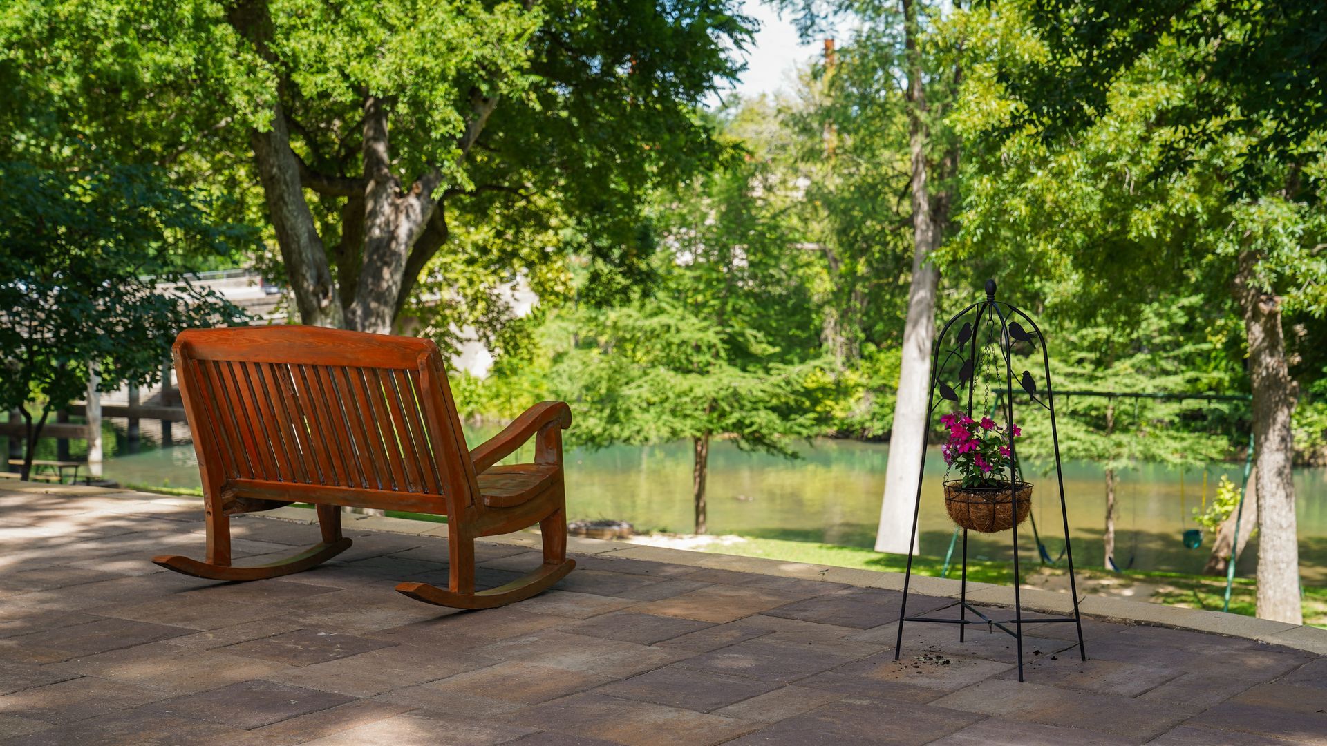Wooden rocking bench on a brick patio overlooking a river, surrounded by green trees and a flower stand.