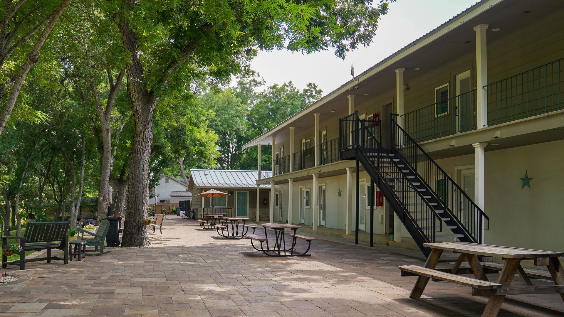 Exterior of a two-story building with a brick patio, trees, and picnic tables. Black metal staircase on the side.