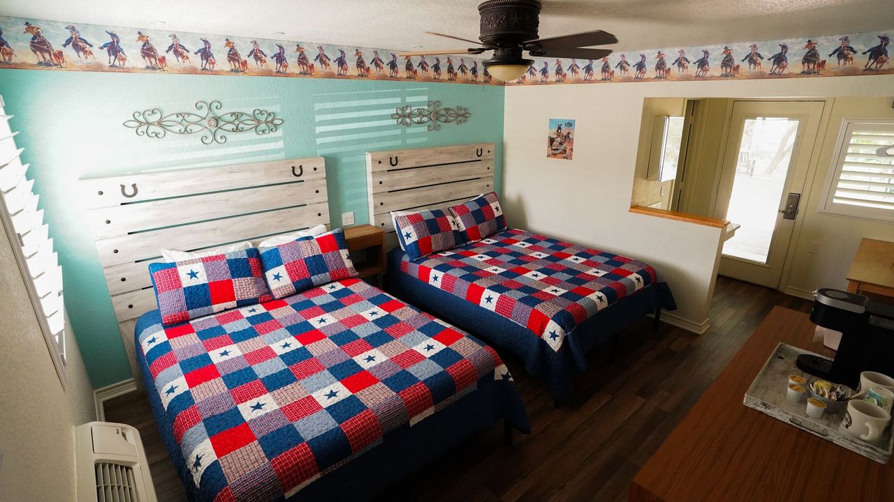 Two beds with red, white, and blue patchwork quilts in a Western-themed motel room with decorative wallpaper.