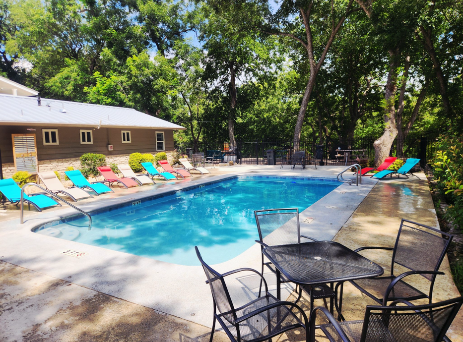 Pool with blue water surrounded by lounge chairs. Metal table and chairs in foreground. Trees and a building in the background.