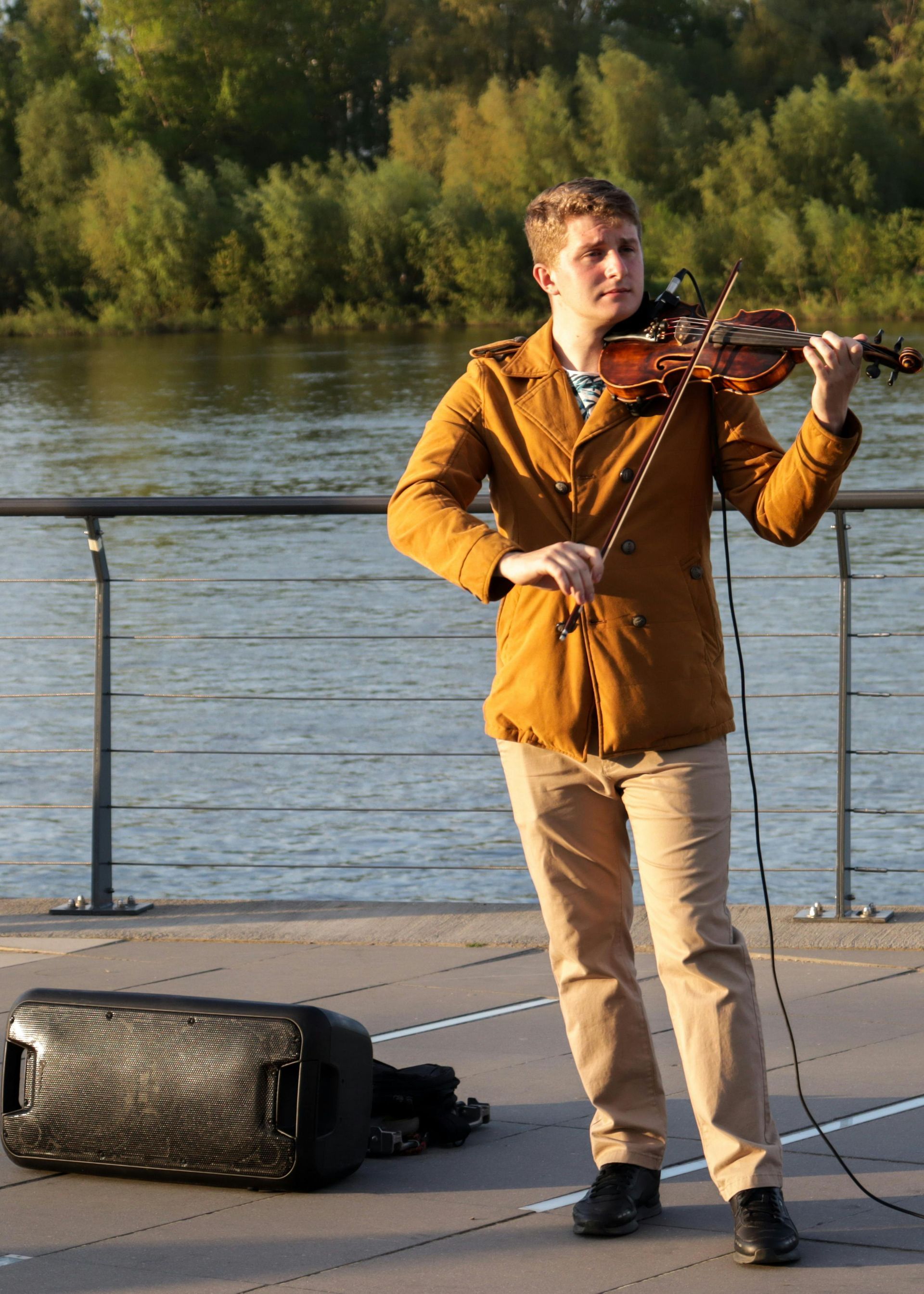Man playing violin outdoors near water, wearing tan jacket and pants.